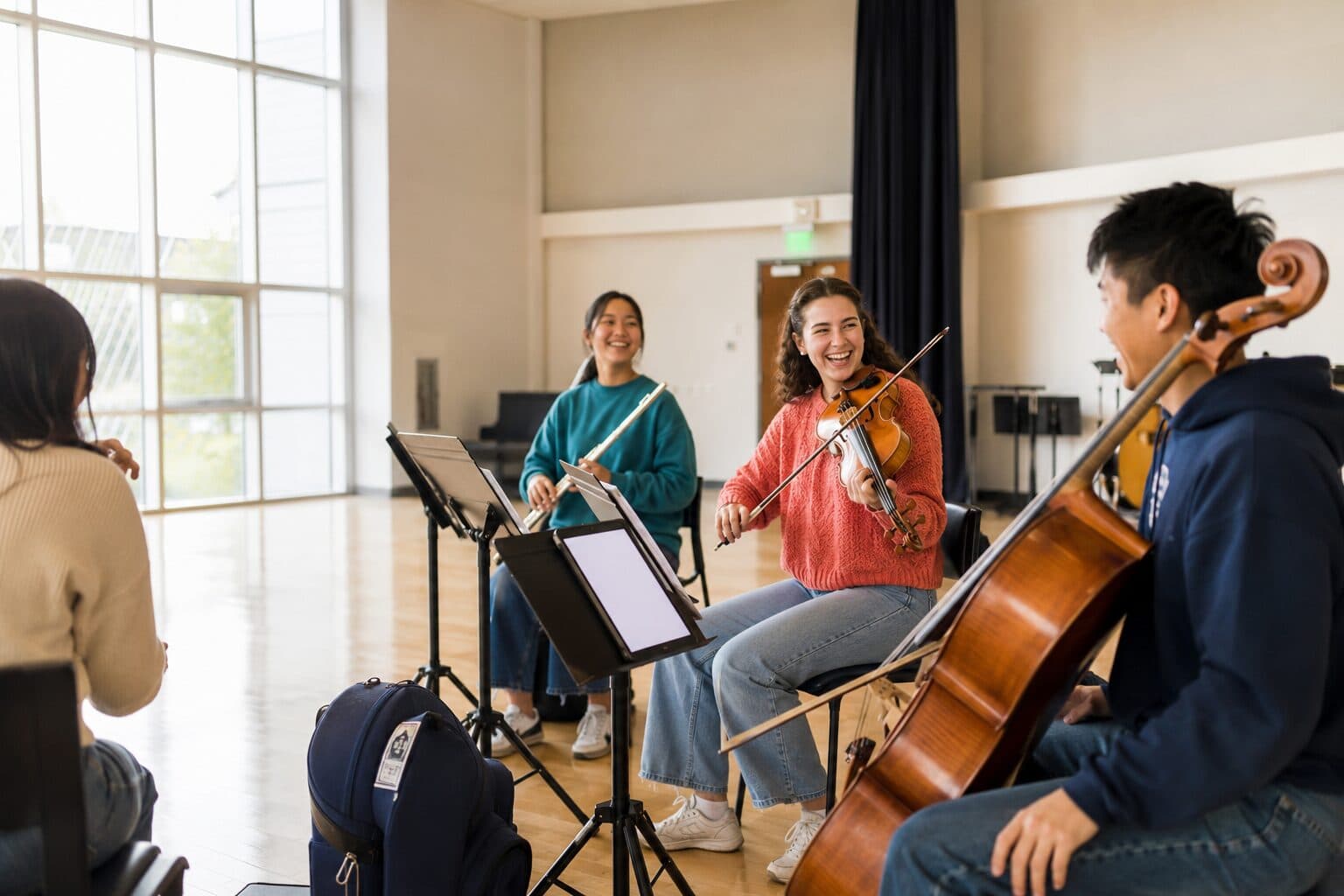 A flautist, a violinist, and a cellist rehearsing together in a bright hall, laughing between phrases, with a tablet on the stand.