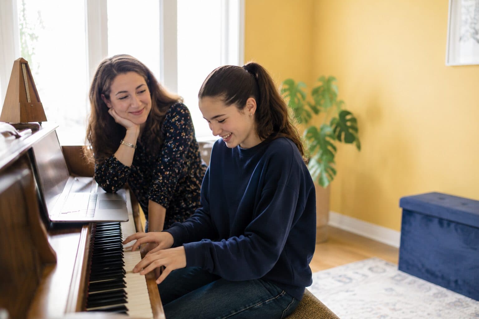 A teacher sitting beside a student at an upright piano, both looking at the keys as the student plays.