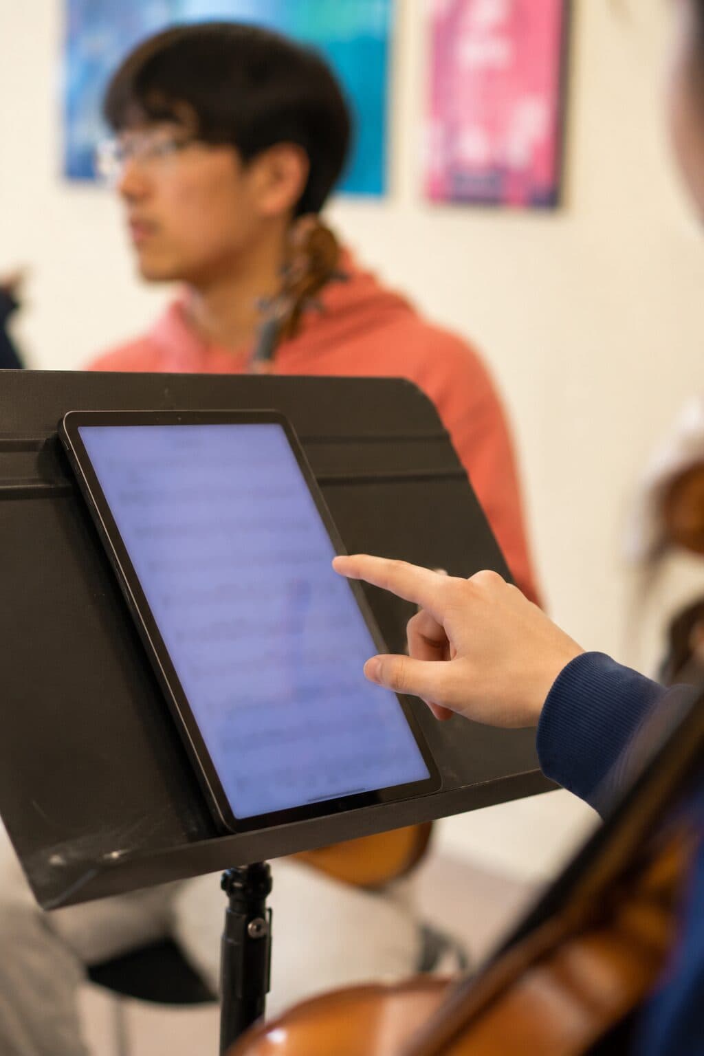 A musician's hand reaching to a score displayed on a tablet propped on a music stand, a cellist just out of focus behind it.